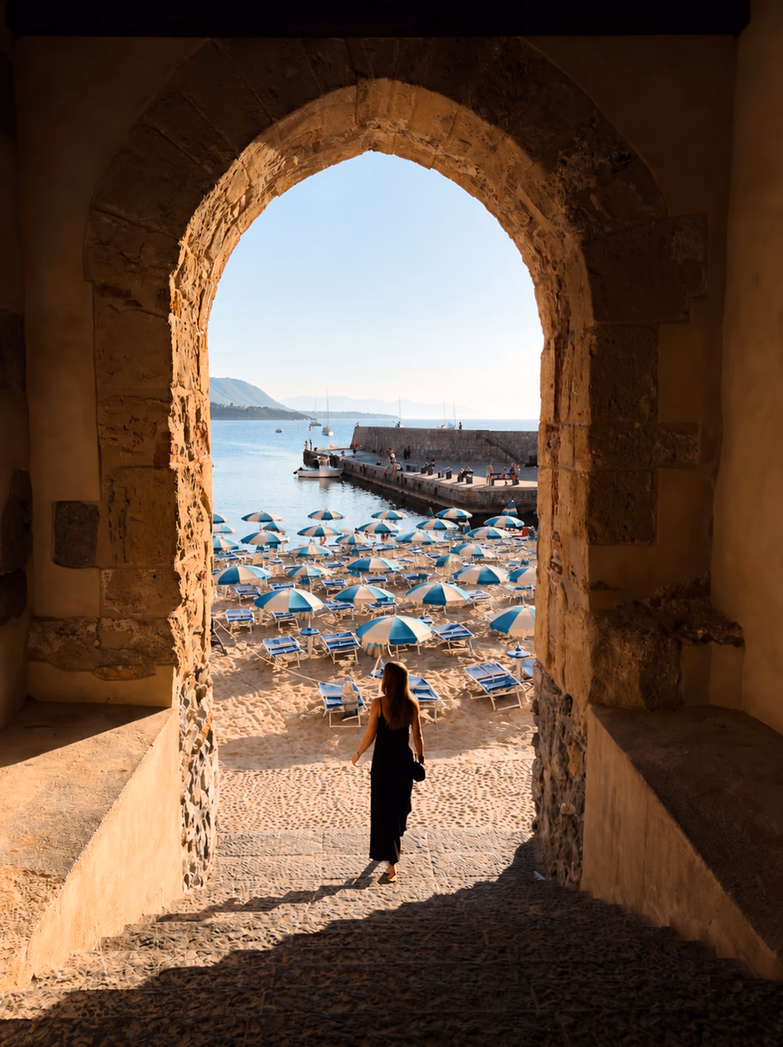 Cefalù beach viewed through a warm stone arch, with a traveller photographed from behind